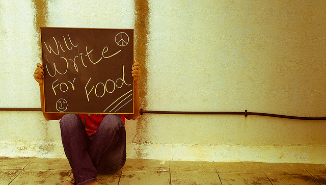 A person sitting on a tiled floor holding up a placard that reads WILL WRITE FOR FOOD.