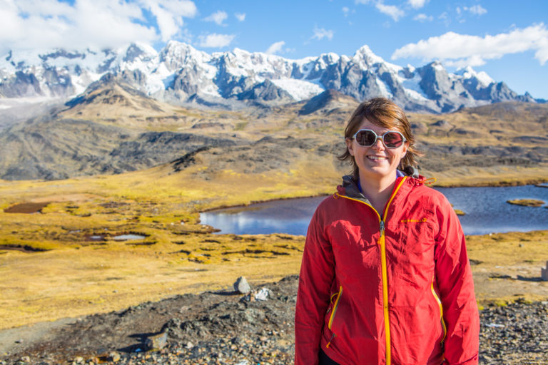 Lizzie Wade in large sunglasses and a red walking jacket with yellow trim smiles at the camera. In the background is a range of snow-topped mountains with flatter grasslands and a small lake just behind her.