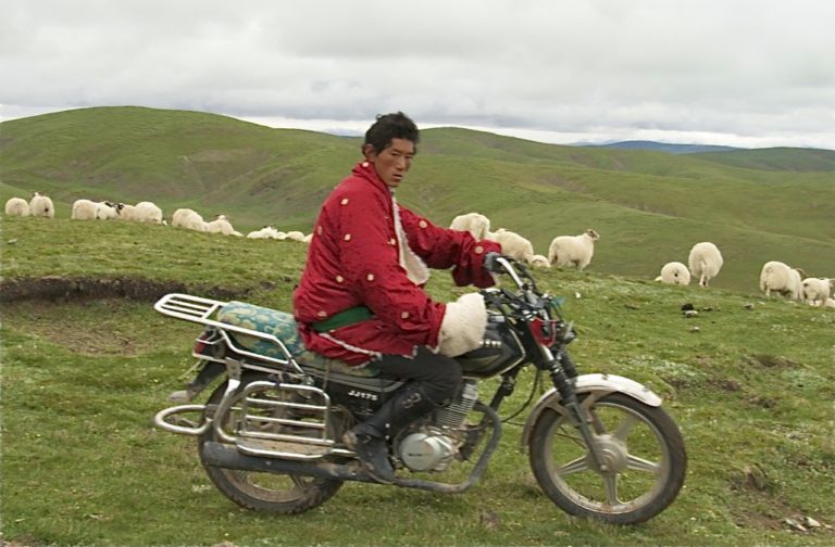 A young man in a bright red coat with gold spots, green belt and large woolly cuffs and collar is on an old motorbike, looking at the camera. Sheep and green hills are in the background.