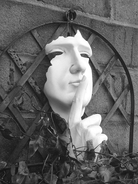 A black and white photograph of a garden trellis with leaves and a white sculpture of a partial human face with the right index finger held up to the lips.