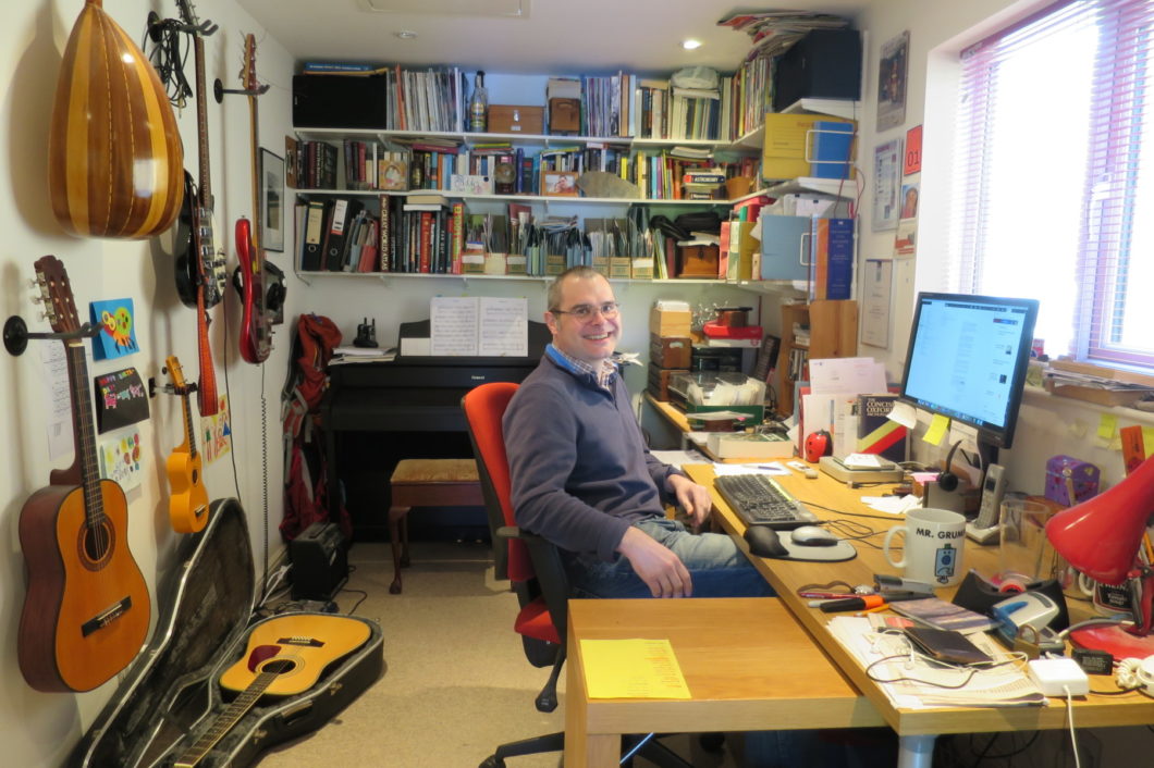 Mark Peplow in his office, sitting at his desk. A bookshelf and a large assortment of musical instruments are nearby.