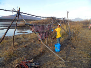A smiling child who is hanging fish on a wooden structure. There is water nearby and mountains in the background.