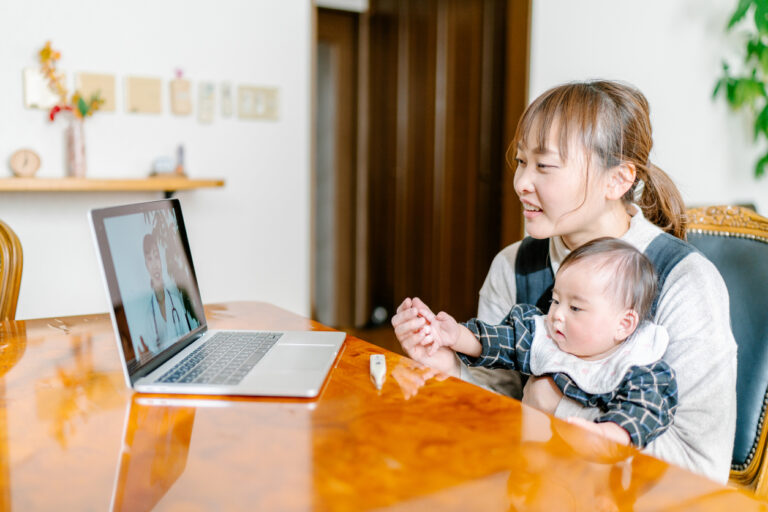 A woman with an infant on her lap sits at a table having a video call with a physican.
