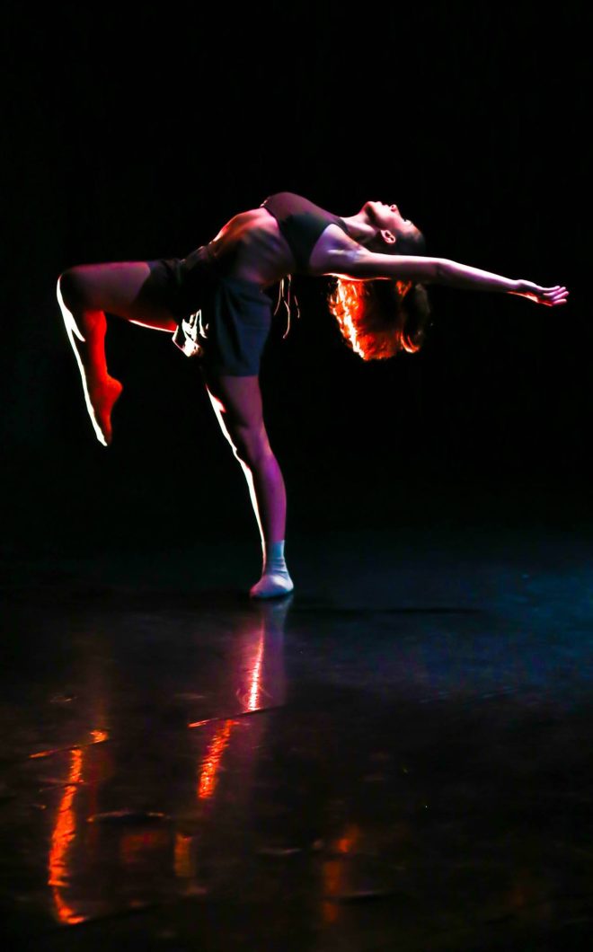 A woman on a stage lit by a warm light dances, folding her body backwards, lifting her knee up and extending her arms above her head.