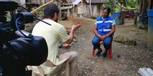 A man and woman sit across each other on an unpaved street surrounded by wooden homes. Behind the man, a camera points in the direction of the woman.