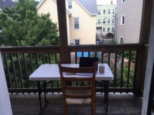 A wooden chair and a white desk with a mug next to open computer on it on a balcony.