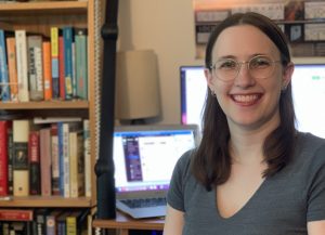 A woman sits smiling in front of a computer screen.