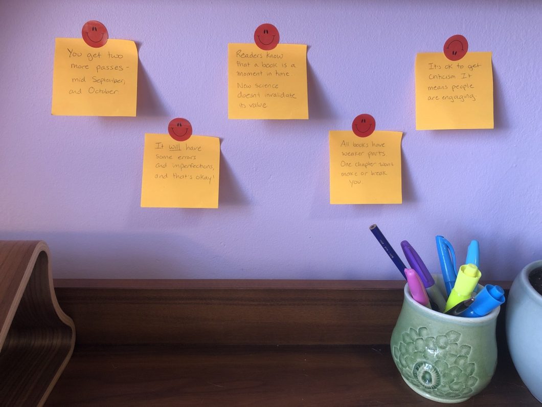 Five orange post-it notes stuck to a purple wall behind a desk, with affirming messages on them.