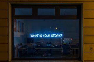 A window with the words WHAT IS YOUR STORY written in neon blue on the glass.