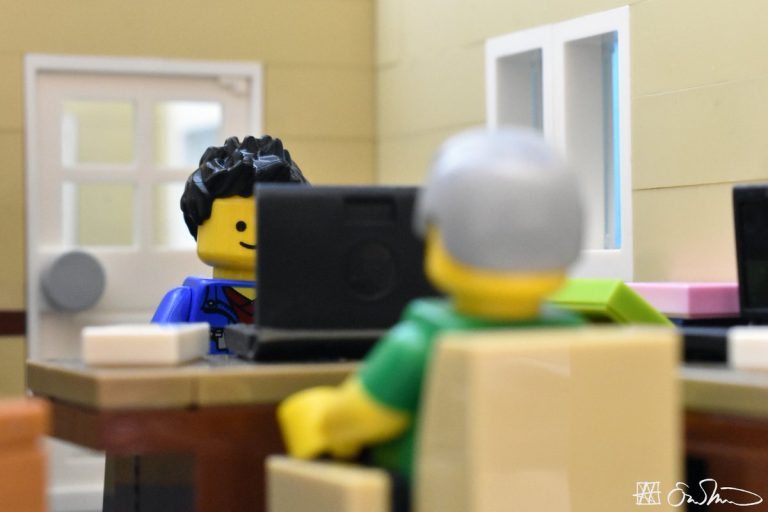 A Lego graduate student sits at a desk behind a black laptop computer. His advisor watches him from the opposite side of the desk.