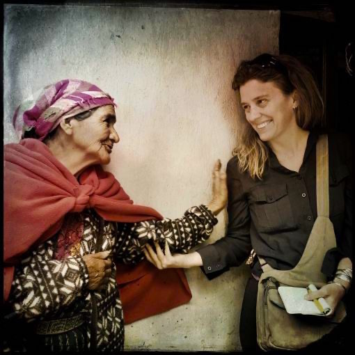 Two women smiling at each other while leaning against a wall and grasping each other's arms.