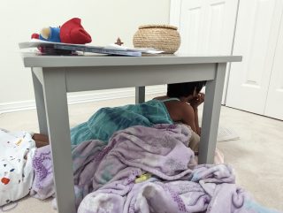 A child snuggles up with a purple blanket under a table and reads a book.
