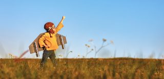 A child wearing cardboard airplane wings and a cloth helmet and goggles stands in a field with one arm thrust upward, as if preparing for flight.