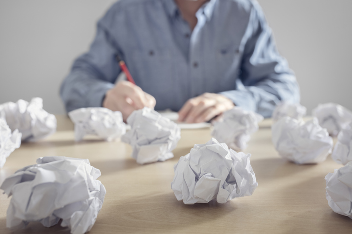 A bunch of crumpled paper balls sit on a desk in the foreground. In the background, a writer sits at the desk with pencil and paper, tyring again.