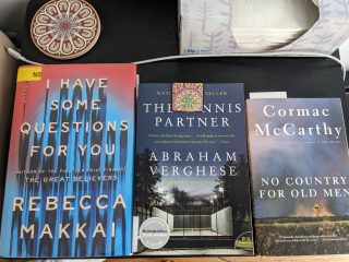 A closeup of three books—I Have Some Questions for You, by Rebecca Makkai, The Tennis Partner, by Abraham Verghese, and No Country for Old Men, by Cormac McCarthy—sitting on a nightstand.