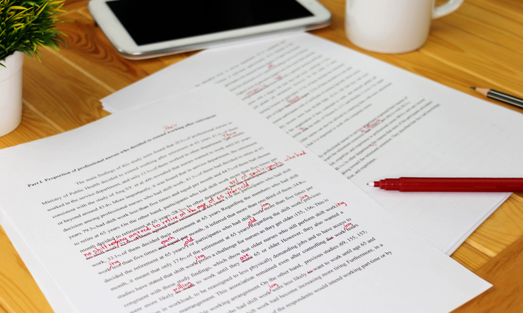 Pages of text edited with a red pen sit on a desk.
