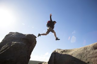 A person wearing a backpack leaps over a chasm between two large rocks.