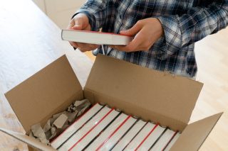 An author opens a box full of hardcover copies of their new book and looks at the cover of one of them.