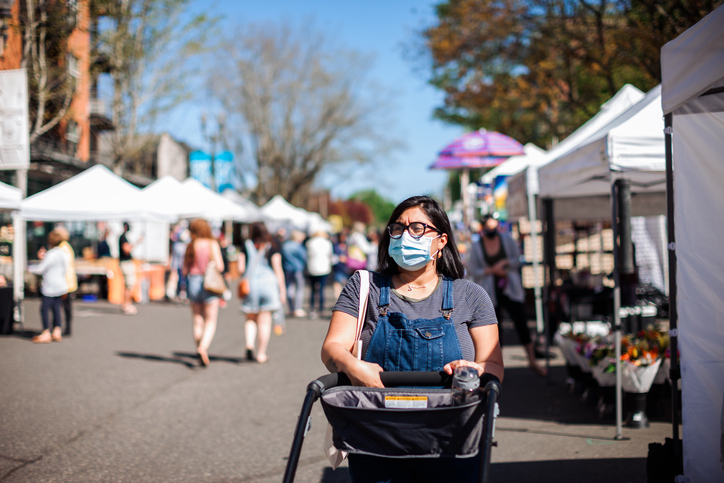 A woman wearing a mask pushes a stroller through an outdoor market.
