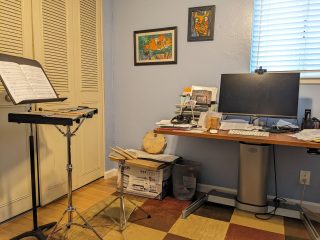 An office with light-blue walls, a standing desk holding a large computer screen, and a glockenspiel with sheet music.