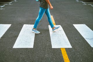 The lower half of a person in blue jeans is seen walking in a crosswalk in a street.