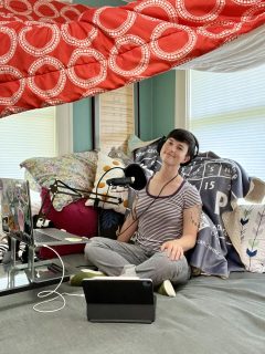 Ari siting on their daybed recording studio behind two screens and a microphone.