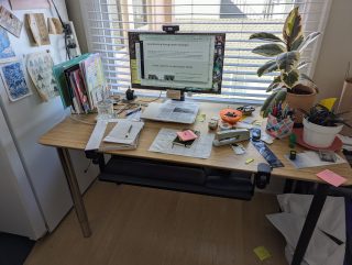 A desk holding a computer and various office supplies sits next to a refrigerator. 