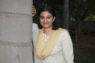 A woman standing next to a pillar, wearing a cream-colored dress and a gold scarf.