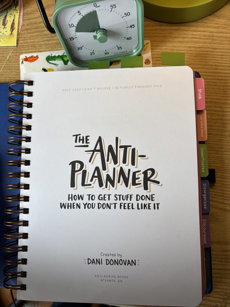 A white spiral-bound notebook labeled The Anti-Planner (created by Dani Donovan), with a small green analogue timer nearby.