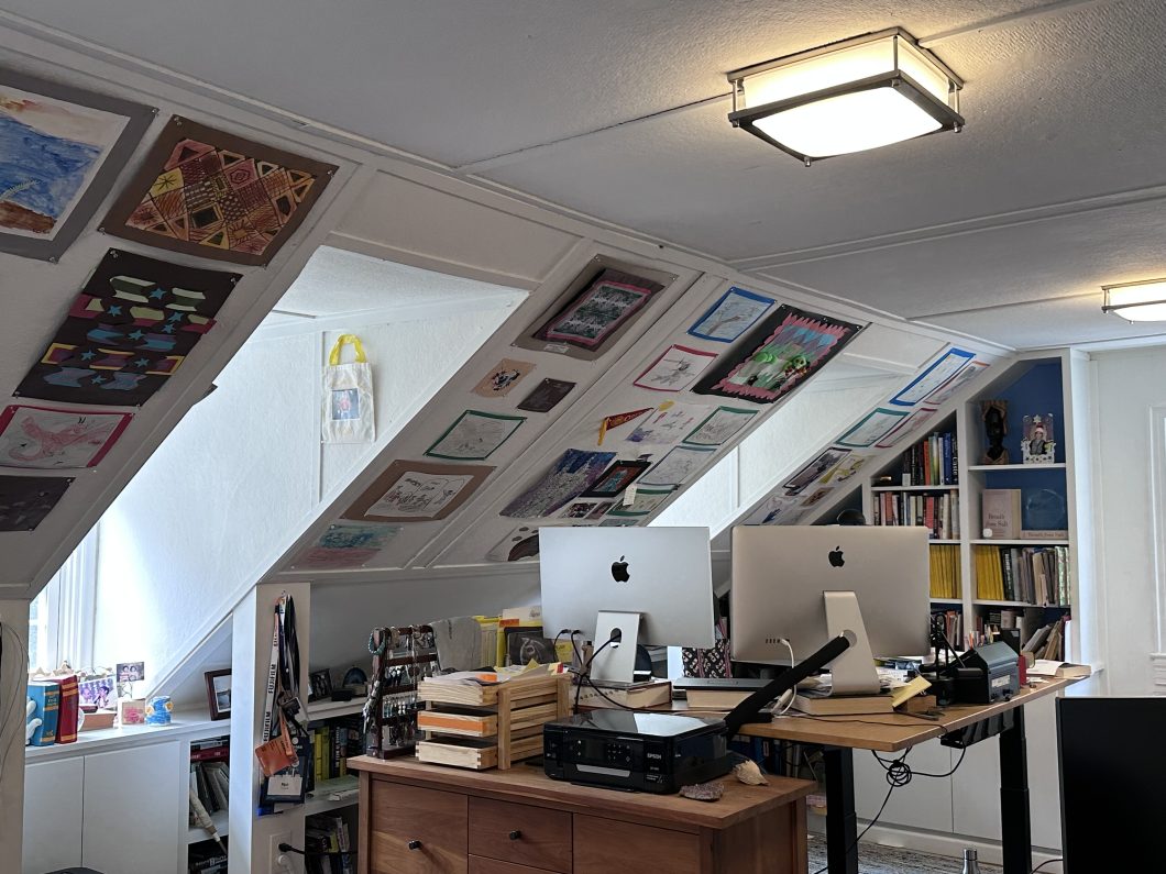An attic office with two windows, two computer monitors, and a sloped wall covered in artwork. 