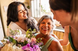 Young sisters giving flowers to grandmother in a sunny room. All of them are smiling.