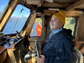 A smiling Boris Hernández stands at the helm of his boat wearing a navy blue coat and a squash-colored winter hat.