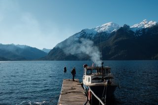 A person stands at the end of a dock next to a boat. I the background there are snow-capped mountains.