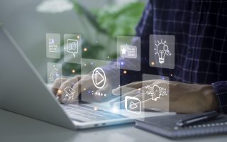 A closeup of a man's hands working on a laptop computer. A notebook sits on the desk next to them. Between the computer and the man there are nine imaginary, holographic windows representing various computer functions.