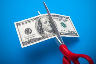 Red-handled scissors cut through a 100-dollar bill in front of a sky-blue background.