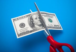Red-handled scissors cut through a 100-dollar bill in front of a sky-blue background.