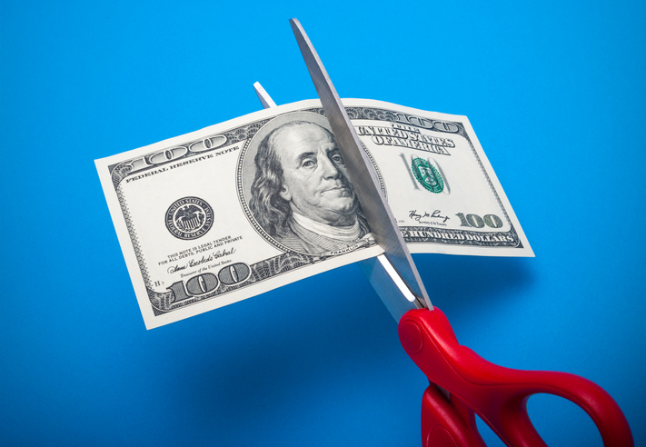 Red-handled scissors cut through a 100-dollar bill in front of a sky-blue background.
