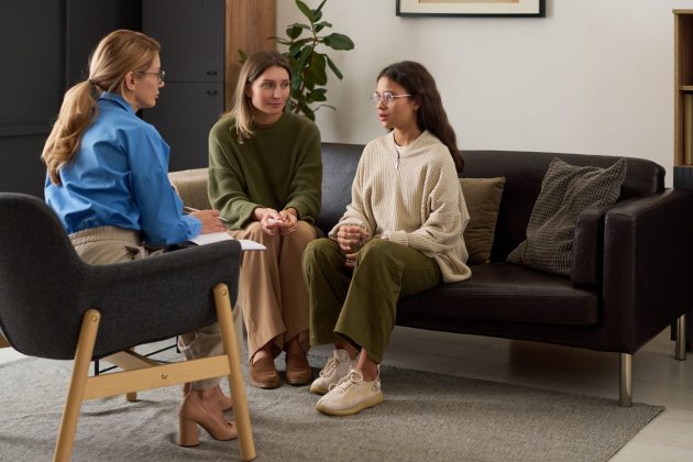Young girl and a female adult sit on a couch. They are speaking to a woman sitting in a chair who is taking notes.