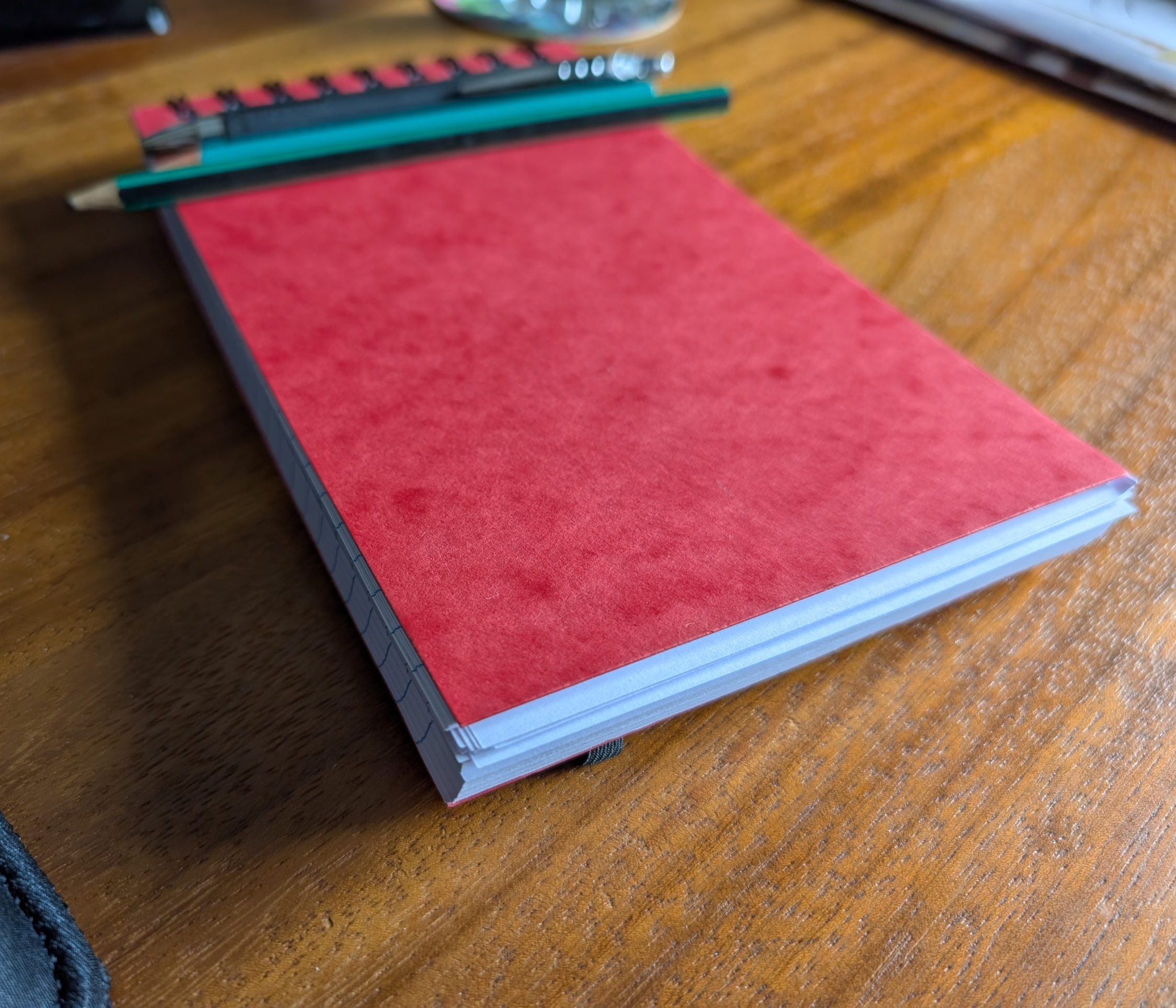A red reporter's notebook sitting on a wooden desk, with pencils and pens resting on top of it.