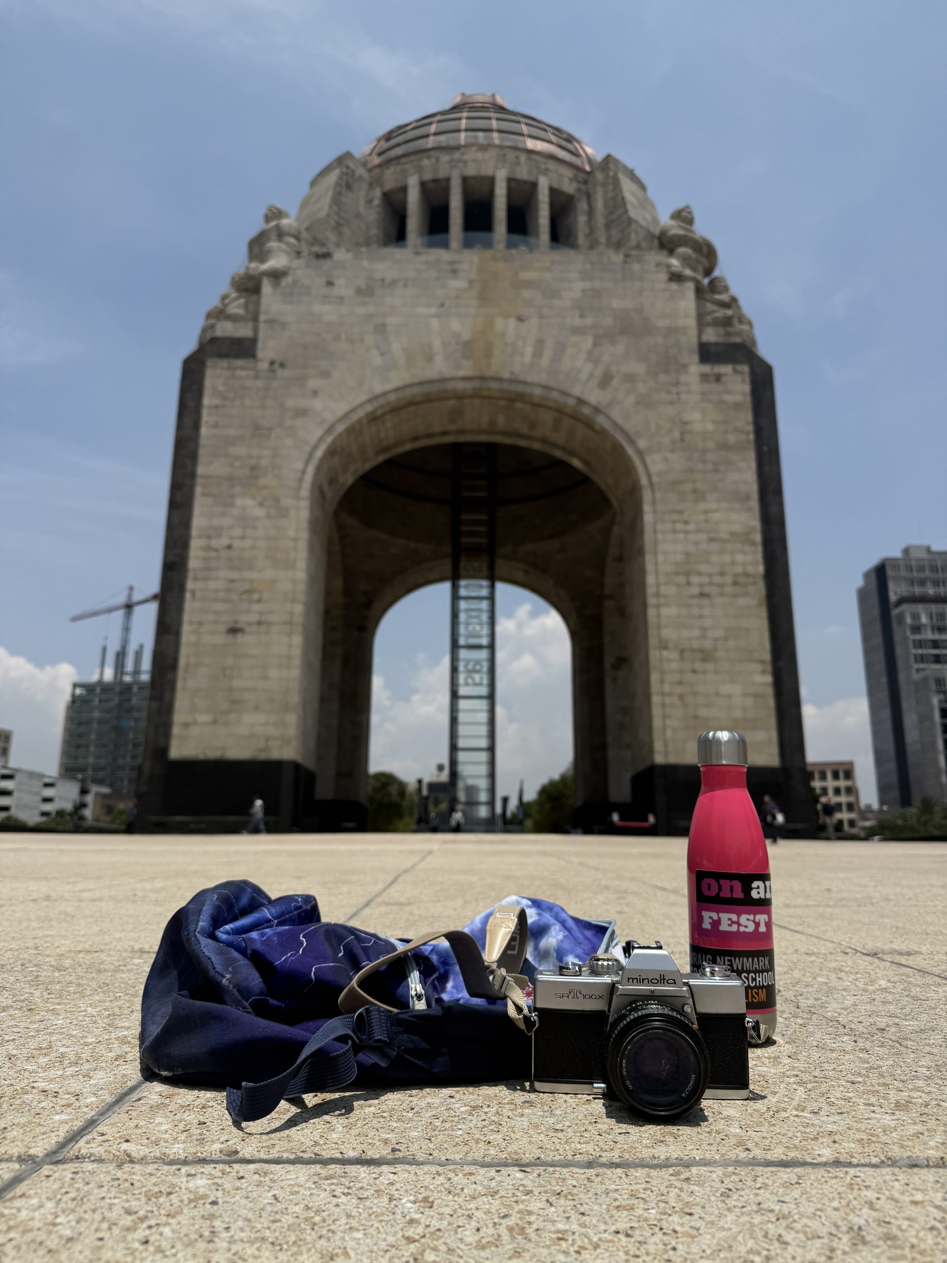 A backpack, camera, and water bottle in the foreground, with an arched building in the background