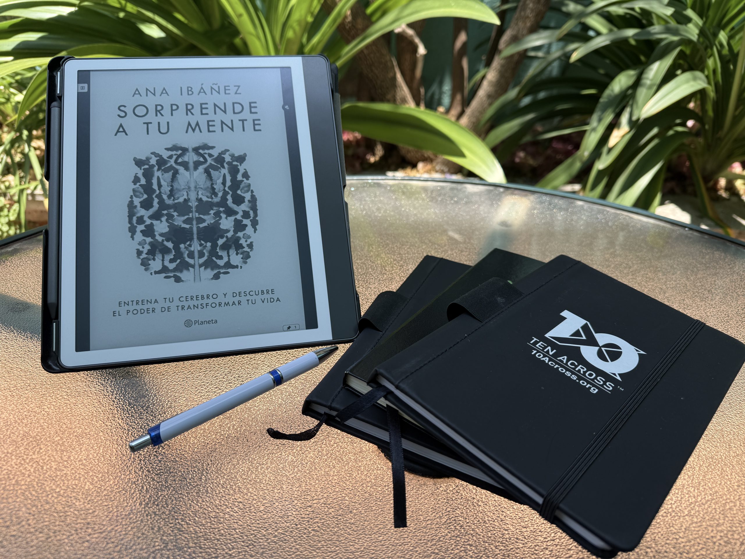 A Kindle Scribe displaying the book Sorprende a Tu Mente by Ana Ibañez, a pen, and three notebooks on a table outside