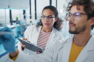Two people in a laboratory setting wearing white lab coats; one wears safety goggles and holds a clipboard. The other person is writing something on a board as the other observes.