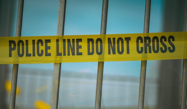 Image of yellow police tape with the words "Police line do not cross" in front of iron bars